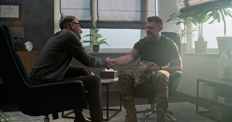 Caucasian military man in uniform sits in chair, talks to psychiatrist and discusses progress after treatment or psychotherapy. Senior psychotherapist smiles, listens to male soldier, asks questions.