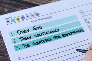 Hand writing Christian priorities in notebook: obey God, pray continually, be grateful to Jesus Christ. Close-up. Biblical concept of spiritual growth, faith, and thanksgiving.