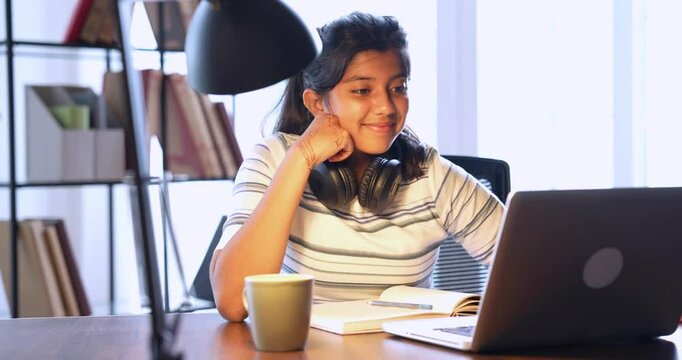 Pretty Indian Asian teenage girl attending an online class from home, studying on a laptop at her study table with headphones on, focused on remote learning in a comfortable home environment