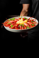 A plate of red boiled crayfish garnished with lemon wedges and herbs, held by a person against a dark background. Perfect for culinary, seafood, and gourmet food themes.