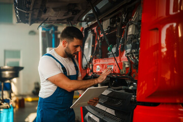 an experienced mechanic inspecting the engine of a red truck in a workshop, holding a tablet