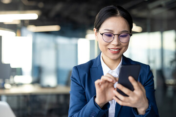 Smiling Asian businesswoman using smartphone in office setting. Confident professional engaged with technology, showcasing productivity and modern communication. Perfect for business, technology