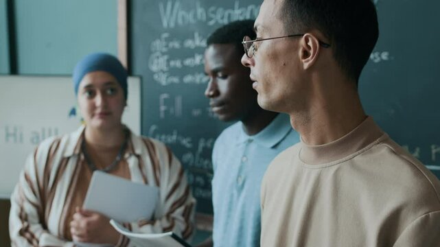 Three multiethnic young adult students with paper sheets in hands making report to Caucasian female student while standing by chalkboard during English lesson