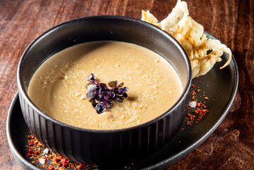 A close-up of a creamy soup in a black bowl garnished with purple flowers, served with crispy bread on the side, on a wooden table sprinkled with spices. Perfect for culinary, food