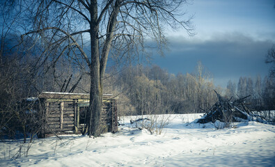 Naklejka premium An old abandoned wooden hut sits quietly on a small country road, surrounded by deep winter snow with a dark and gloomy sky above. The crumbling hut is set against a snow-covered field.