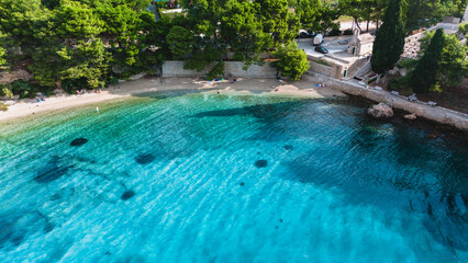 Visitors bask in the vibrant turquoise waters surrounding Brac Island, Croatia, as sunlight sparkles on the calm sea. Lush green trees provide shade along the shoreline, creating a serene atmosphere.