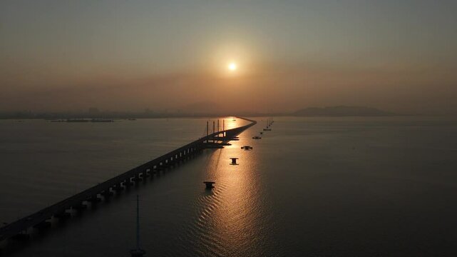 Penang Bridge at sunrise, framed by two TNB monopole electric transmission towers. The golden morning light casts a warm glow over the scene, highlighting the bridge's structure.