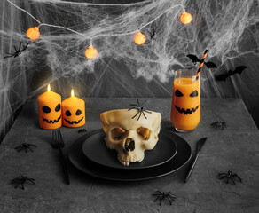 Halloween table setting with skull in black dishes, burning candles, garland on dark grey background with cobwebs.