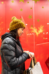 Woman in coat and winter hat checking time in front of beautiful Christmas decorated shop window while shopping in the city