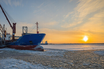 Naklejka premium In winter, in severe frost, a cargo ship stands on a pier for loading against the background of a frozen river and the setting sun