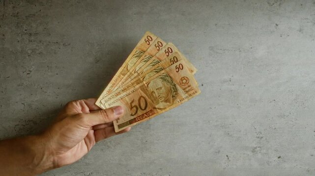 Hand holding brazilian 50 reais bills against a plain concrete background