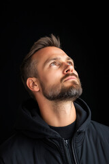 Fototapeta premium A man gazing upward with a thoughtful expression, featuring a beard and wearing a dark hoodie, set against a black background.