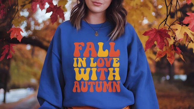 A young woman wearing a blue mockup sweatshirt with fall colors and a themed background for the autumn season.The sweatshirt has a large orange and yellow text that says "Fall in Love with Autumn".