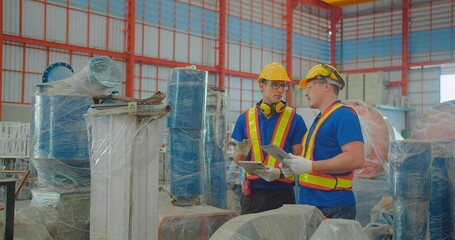 Two workers wearing safety gear inspect materials in a modern warehouse. They collaborate closely,...