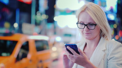 A young woman uses a smartphone on a busy street in Manhattan against the backdrop of bright advertising lights, yellow taxis are passing by - one of the symbols of New York