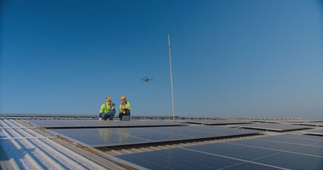 Two engineers with safety vests and hard hats are inspecting solar panels with a drone flying overhead against a clear blue sky