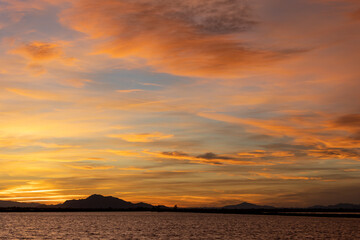 The sky glows with orange, yellow, and red hues as the sun sets over the mountains, reflecting in calm waters below, creating a peaceful and colorful evening scene