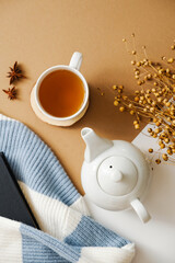 Autumn still life with teapot, cup of tea, dry flowers, knitted sweater with book on brown background. Cozy fall flat lay composition