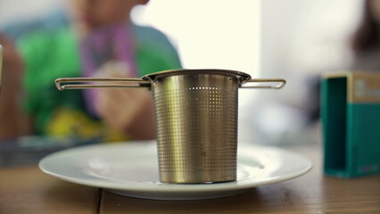 Stainless steel tea infuser placed on a white plate, with a blurred background. the simplicity and utility of everyday kitchen tools in a relaxed home environment