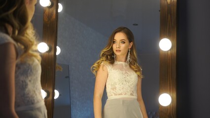Beautiful blonde woman with long wavy hair wearing an elegant white lace dress looking at herself in the mirror © Довидович Михаил