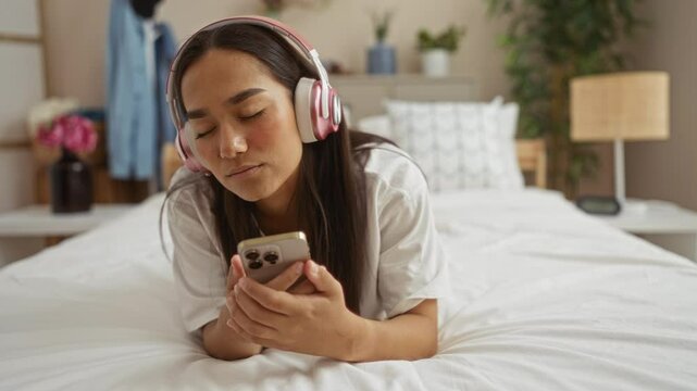 Young woman lying on bed in bedroom listening to music with headphones and holding smartphone, eyes closed, looking relaxed and connected to technology in cozy home interior.