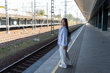 A little teenage girl stands at the train station waiting for the train, she is smiling in a light jacket and pants.