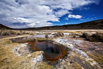 A geothermal hot spring surrounded by grasslands and mountains under a cloudy sky.
