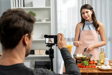 Cameraman recording to woman in chef influencer host cooking spaghetti with meat topped tomato sauce surrounded ingredients recipe, presenting special dish healthy food at modern studio. Postulate.