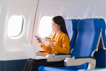 In-Flight Purchases: A young Asian woman enjoys the convenience of online shopping during her flight, using her credit card and smartphone while seated comfortably in an airplane cabin. 