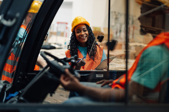 African american female worker smiles at forklift driver in warehouse.