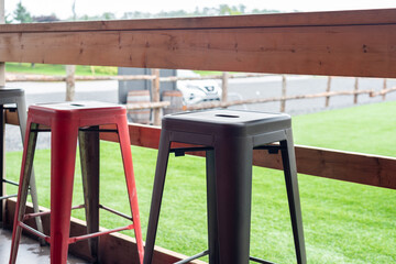 Multiple colorful iron metal stackable bar stools; black, and red colored with footrests. The chairs are on a wooden restaurant patio. There's a lush green fenced in grassy field off the porch.  