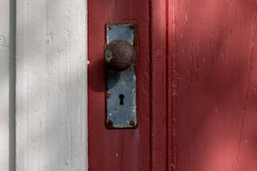 A closeup of a colorful antique red painted exterior wooden door with a rusty metal door knob and steel plate. There's a keyhole in the plate. The trim on the building has wide white weathered boards.