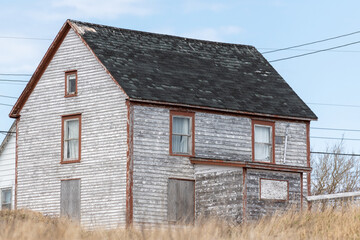 A vintage salt box house that is dilapidated, worn, weathered and abandoned. The exterior is white with red trim. There are multiple double hung windows, black shingles and boarded windows in a field 