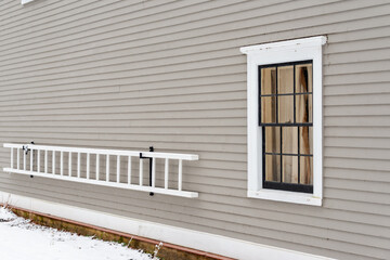 The exterior of a grey colored wooden wall of a house. The vintage building has painted clapboard siding with white trim. A wooden ladder, painted white, covered in ice hangs on hooks on the property.