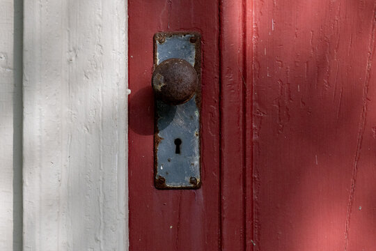 A closeup of a colorful antique red painted exterior wooden door with a rusty metal door knob and steel plate. There's a keyhole in the plate. The trim on the building has wide white weathered boards. - Powered by Adobe