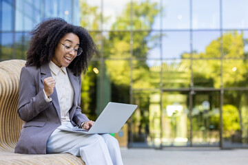 Hispanic businesswoman expressing excitement and success while working on laptop. Seated on bench in front of large glass building. Professional attire, sense of accomplishment and joy.