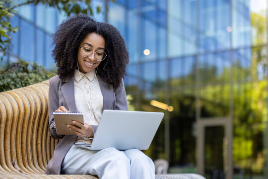 Hispanic businesswoman works outdoors holding notebook and using laptop for online meeting. Shows confidence, professionalism, multitasking in urban environment with glass facade backdrop.