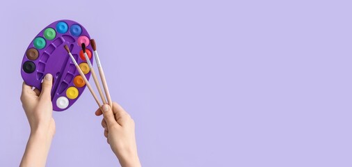Hands of female artist holding brushes and palette on lilac background with space for text