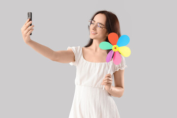 Young woman with toy windmill taking selfie on light background