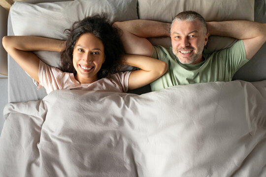 Top view of happy spouses lying in bed covered with blanket and with hands behind heads smiling to camera, above view shot