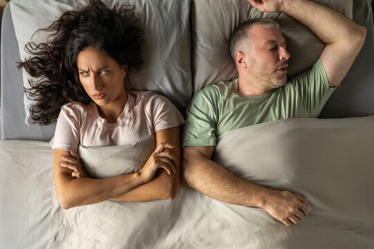 Man asleep, his offended angry wife with crossed arms on her chest lying in bed nearby, above view shot. Relationship problems, quarrel, snoring - Powered by Adobe