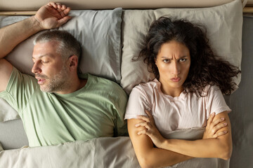 Above view of anger lady with arms crossed lying near snoring husband, unpleasant morning...