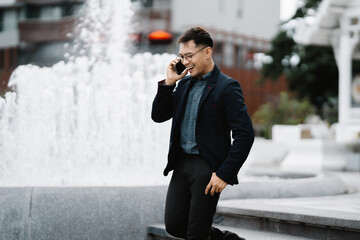 Successful Businessman on Phone Call: A confident and well-dressed businessman in a suit and glasses takes a call in front of a fountain, radiating success and professionalism. The image exudes a sens © Kritdanai