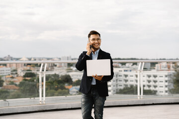 Urban Professional on the Rise: Young Asian businessman, laptop in hand, enjoys a successful phone...