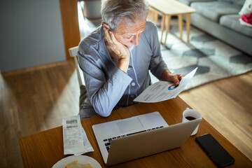 Senior man reviewing financial documents at home with laptop