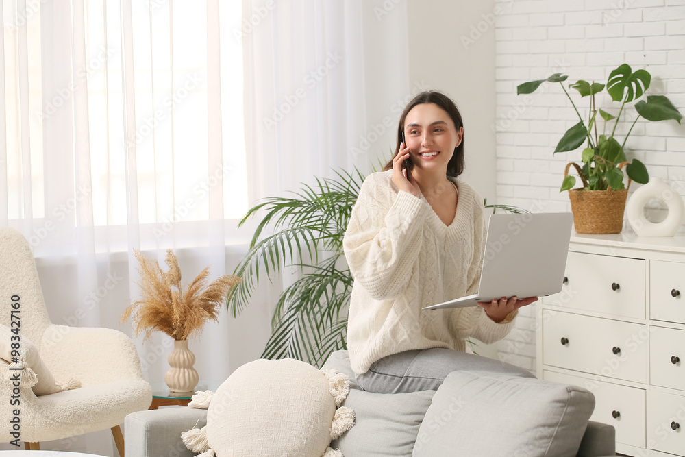 Canvas Prints Young woman with laptop talking by mobile phone at home