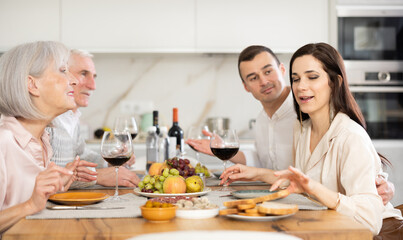 Mature couple are sitting at table, chatting sweetly, discussing and telling news, sharing plans with adult children. Senior woman and man celebrate housewarming party with middle-aged guests spouses