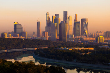 The skyline of downtown Moscow showcases towering modern skyscrapers of business district illuminated by the warm glow of sunset. The river reflects the vibrant colors, enhancing the urban landscape.