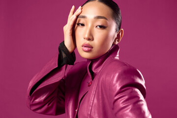 Fashion asian brunette in purple leather jacket and black shirt posing against purple background with hands on head, looking at camera