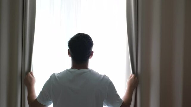 Man stands at window and opens curtains back view.